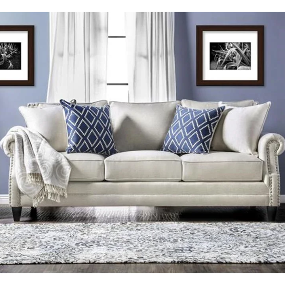 Beige tufted linen sofa with rolled arms, nailhead trim, tapered wooden legs, and accent pillows in transitional style.