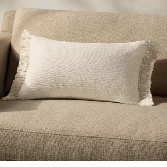 Cream lumbar throw pillow with soft fringe trim, rectangular 13x21 design, made from neutral cotton blend fabric, styled on a sofa for cozy, modern home decor.