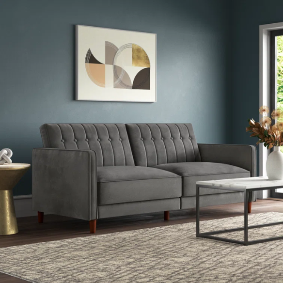 Dark grey velvet convertible sofa with vertical tufting, split-back design, square arms, and brown tapered legs, convertible into a twin-size sleeper.