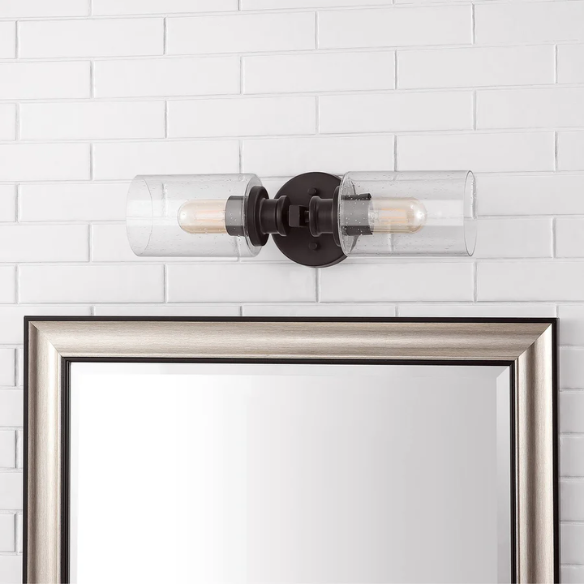 Oil-rubbed bronze wall sconce with dual clear seeded glass cylinder shades and Edison-style LED bulbs, perfect for rustic, farmhouse, or traditional interiors