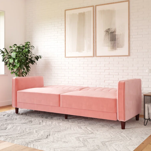 Pink velvet convertible sofa with tufted split-back design, square arms, and tapered brown wood legs, easily converting into a twin-size sleeper.