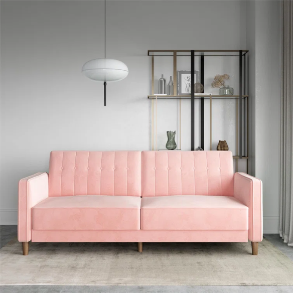 Pink velvet convertible sofa with tufted split-back design, square arms, and tapered brown wood legs, easily converting into a twin-size sleeper.