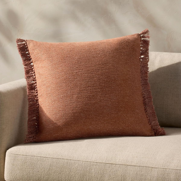 Rust cotton blend throw pillow with fringe, 22x22 square decorative cushion styled on a neutral-toned sofa for cozy, modern home décor.