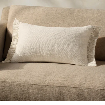 Cream lumbar throw pillow with soft fringe trim, rectangular 13x21 design, made from neutral cotton blend fabric, styled on a sofa for cozy, modern home decor.

